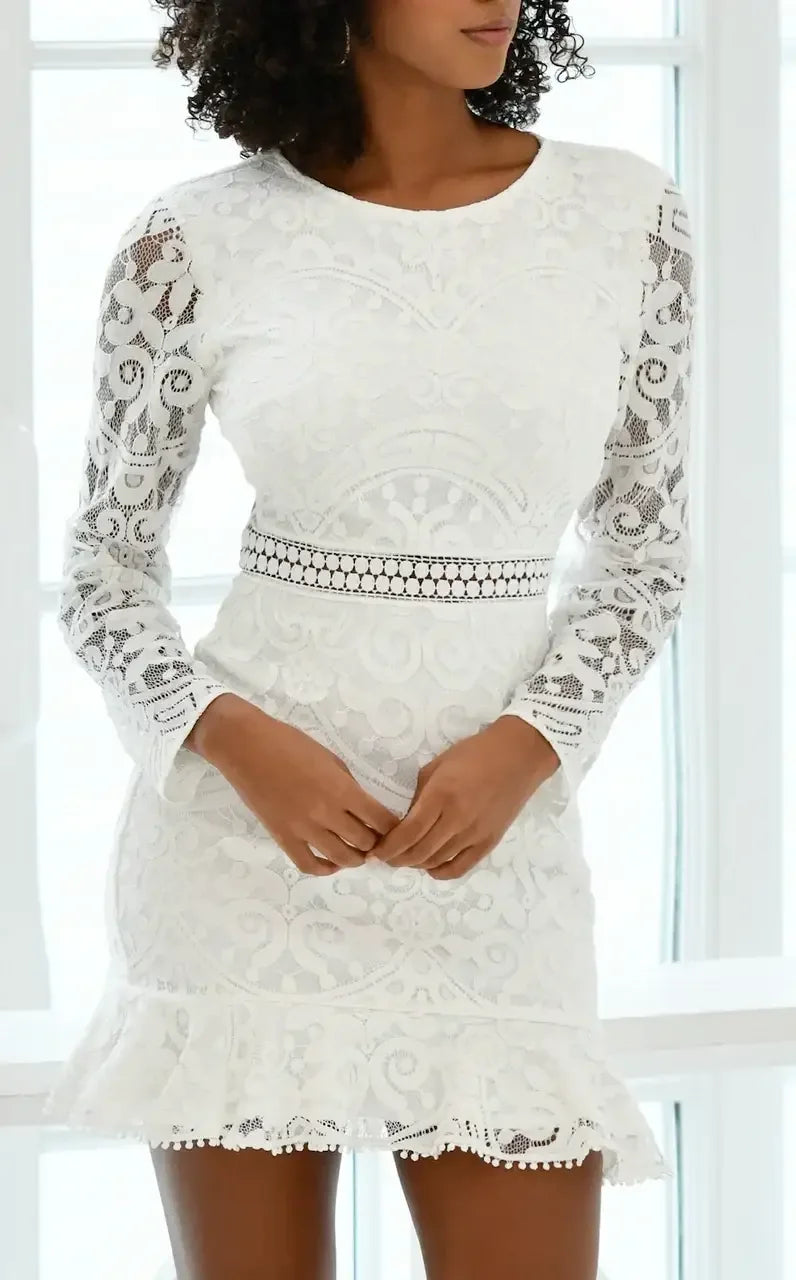 A woman in a white lace dress stands with her hands gently clasped together. She is positioned near large windows, allowing natural light to illuminate the intricate patterns of her dress.