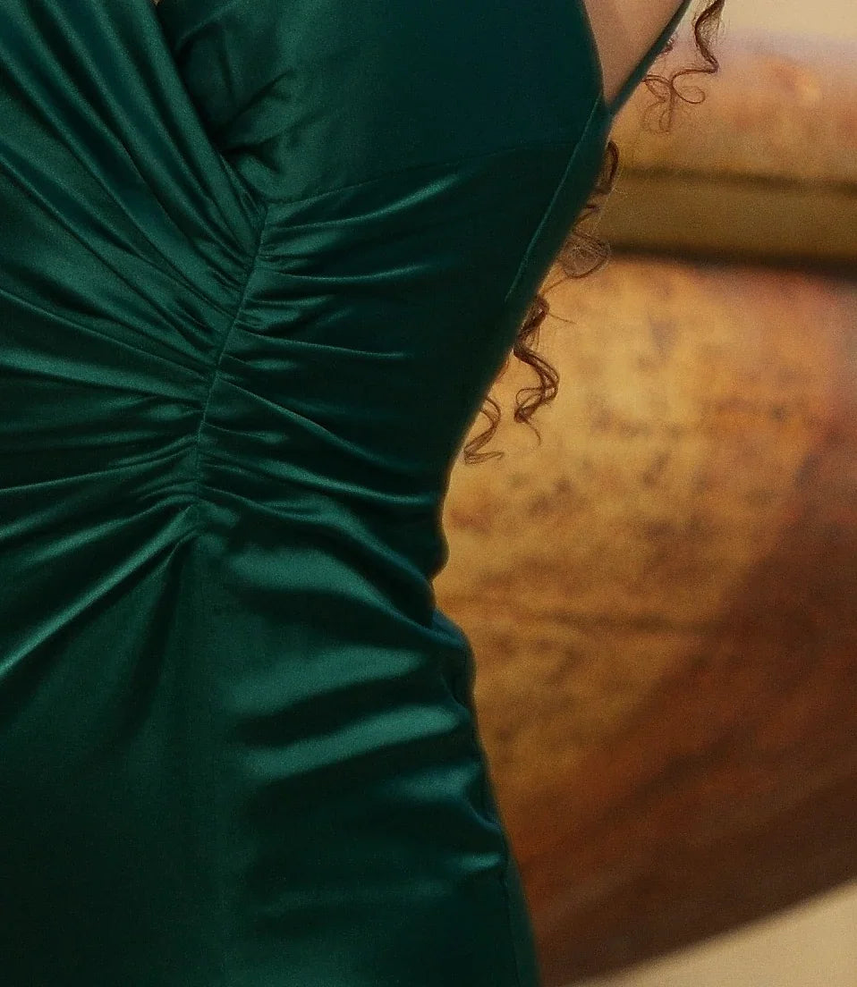 A green, form-fitting dress with ruching details is worn by a figure, who is partially visible. The background features a blurred, warm-toned, textured surface.