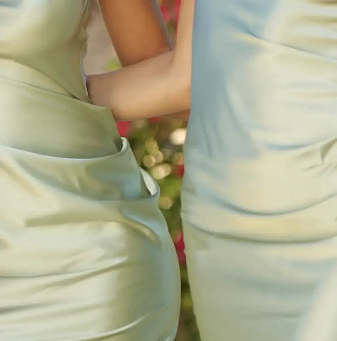 Two individuals are standing close, wearing satin dresses in soft green tones. One person's arm is resting gently on the other’s waist, with a blurred floral background adding color.