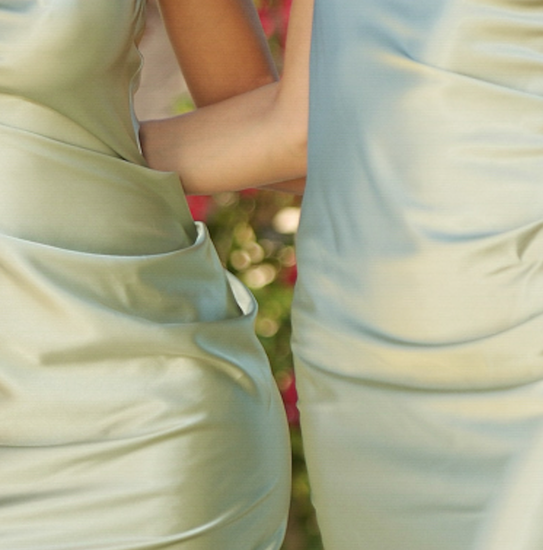 Two individuals are standing close, wearing satin dresses in soft green tones. One person's arm is resting gently on the other’s waist, with a blurred floral background adding color.
