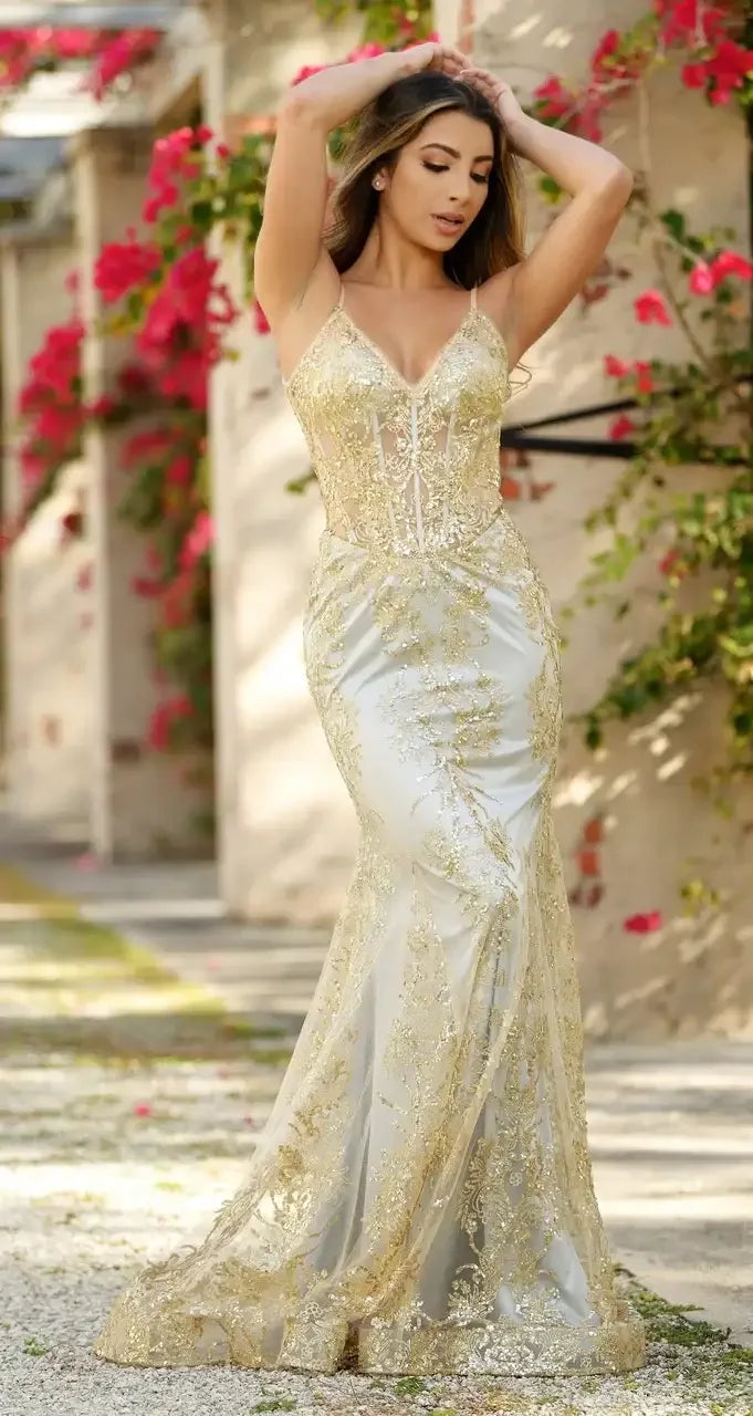 A woman in a shimmering gold and silver gown poses gracefully, her hands raised in her hair, surrounded by vibrant pink flowers and a soft, sunlit outdoor pathway.