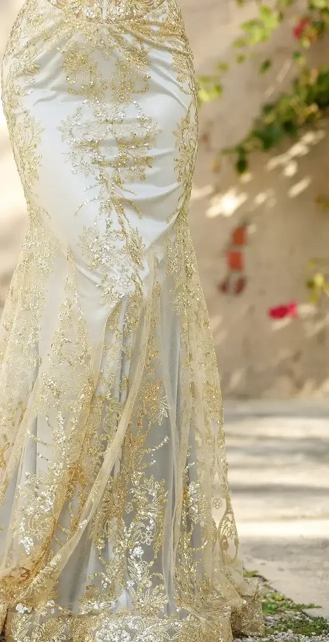 A shimmering, white gown adorned with intricate gold embroidery flows elegantly to the ground, set against a softly lit, blurred outdoor background featuring greenery and floral accents.