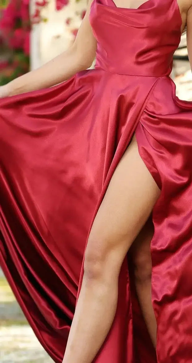 A red satin gown flows elegantly, with a high slit revealing a leg. The backdrop features soft, blurred greenery and hints of pink flowers, creating a vibrant yet serene atmosphere.