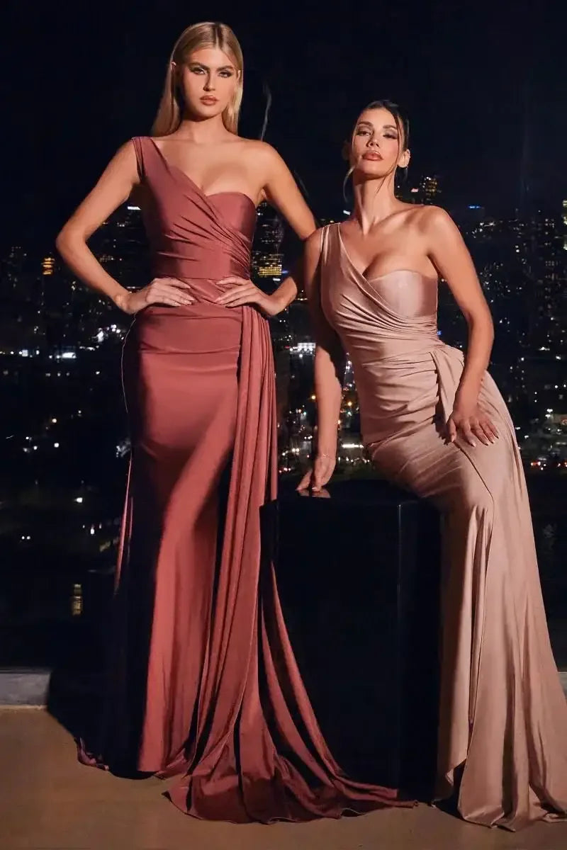 Two women in elegant evening gowns stand confidently against a nighttime city backdrop. The left dress is rich maroon, while the right gown is a soft beige.