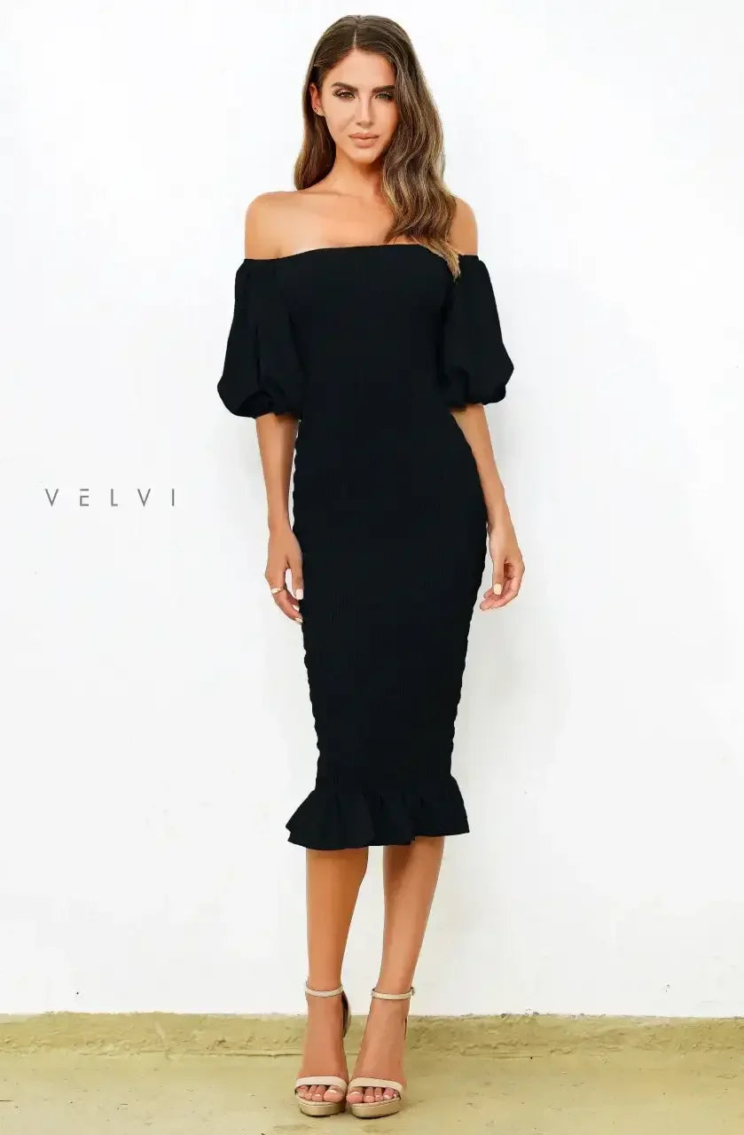 A woman stands confidently in a fitted black dress with off-the-shoulder sleeves, accentuated by a ruffled hem. She wears beige high-heeled sandals and poses against a textured white wall.