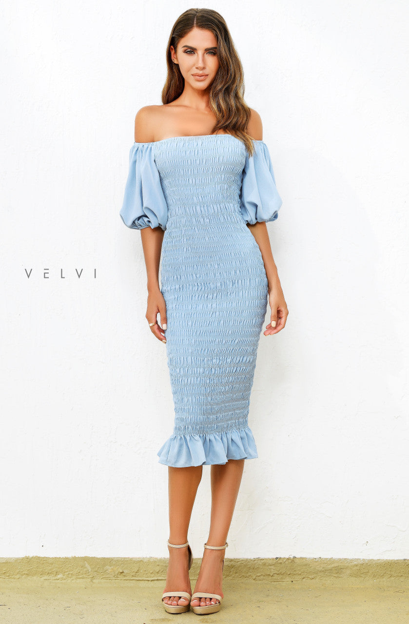 A woman stands confidently in a light blue, off-shoulder, ruched dress with puff sleeves and a ruffled hem. She wears strappy nude heels, set against a white wall backdrop.