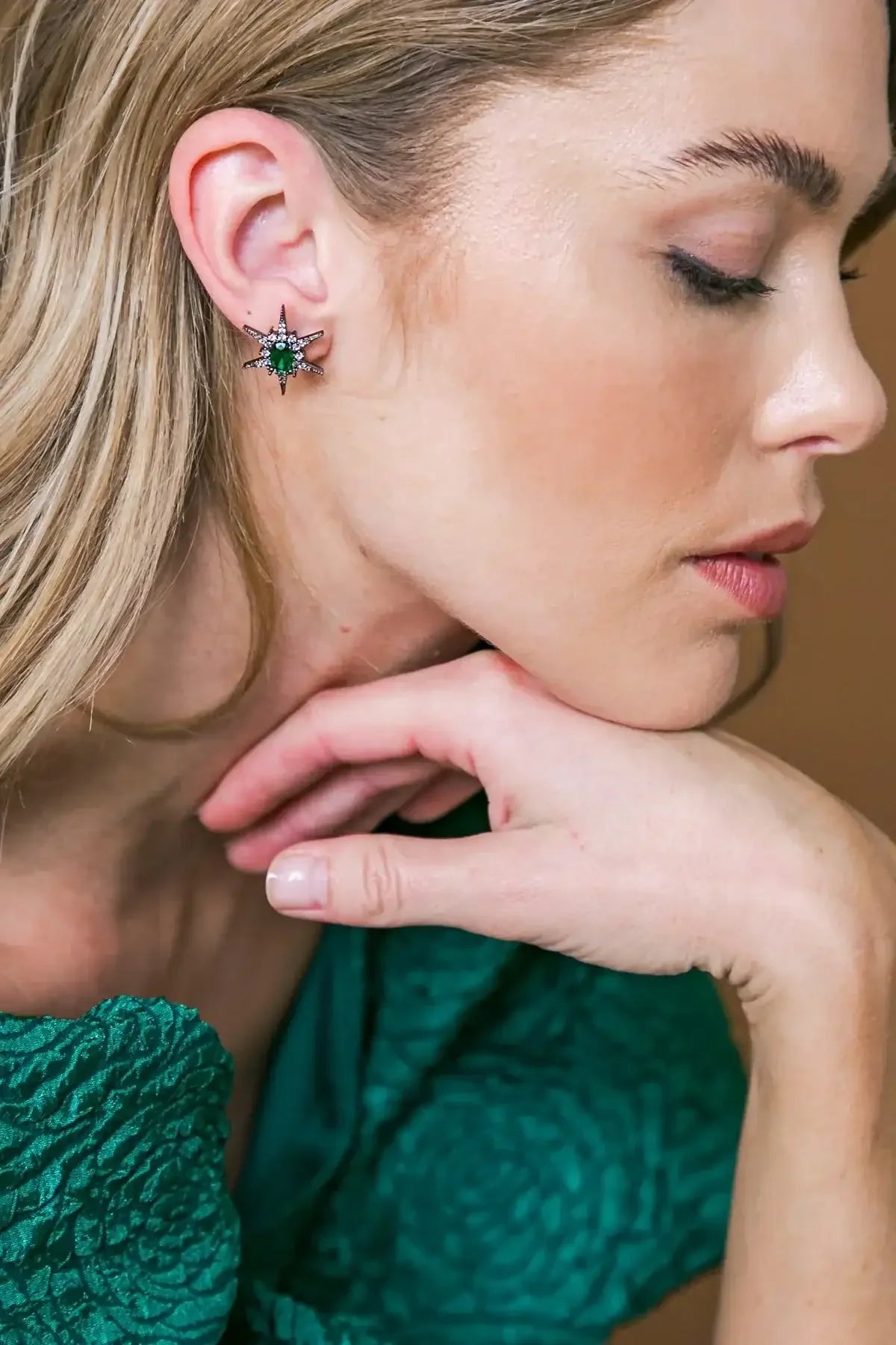 A woman with long blonde hair wears an ornate star-shaped earring with a green gem. She rests her chin on her hand, posed against a neutral background.