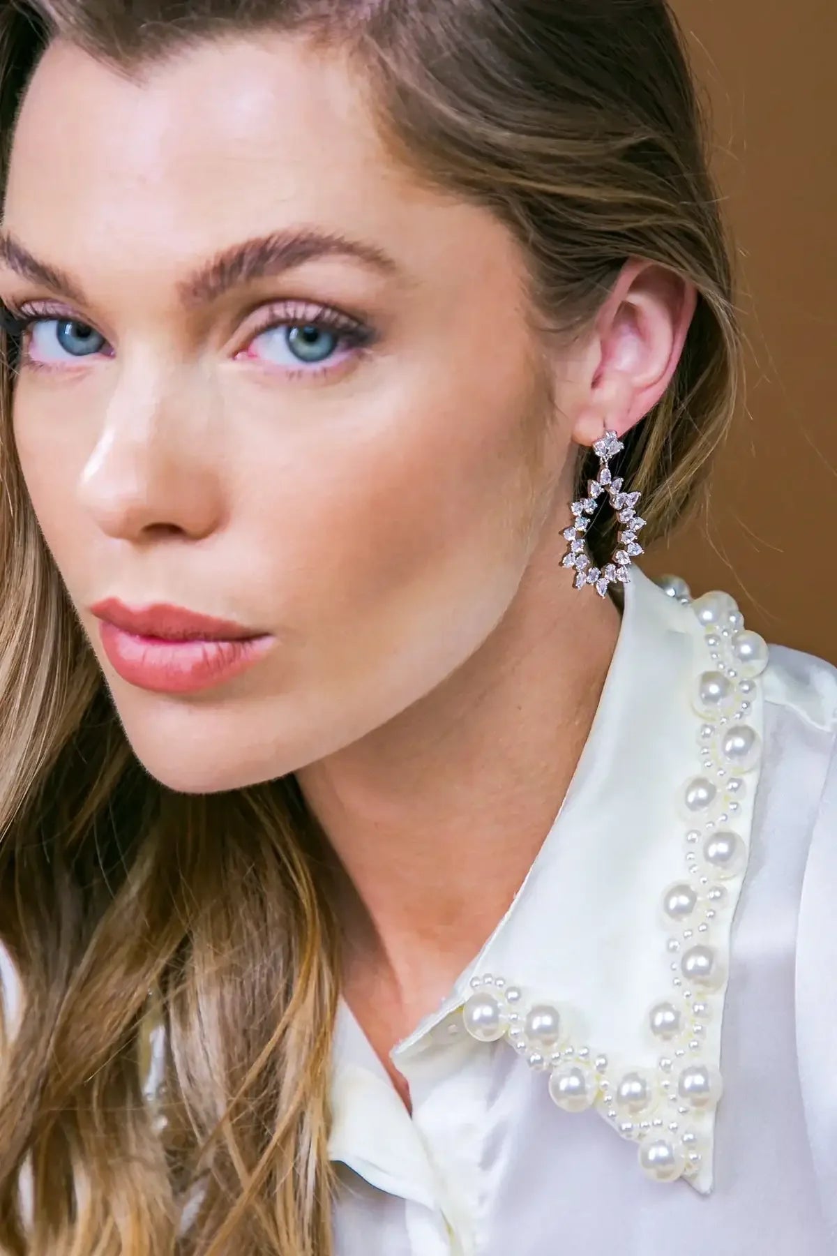 A woman with long hair wears an ornate earrings and a pearl-adorned blouse. She gazes directly at the viewer, set against a simple brown backdrop.