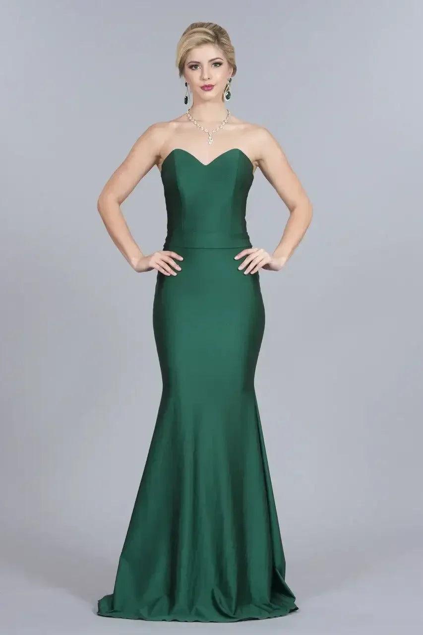A woman stands confidently, wearing a fitted green evening gown that flares at the hem. She poses with hands on her hips against a plain gray backdrop, showcasing her elegant jewelry.