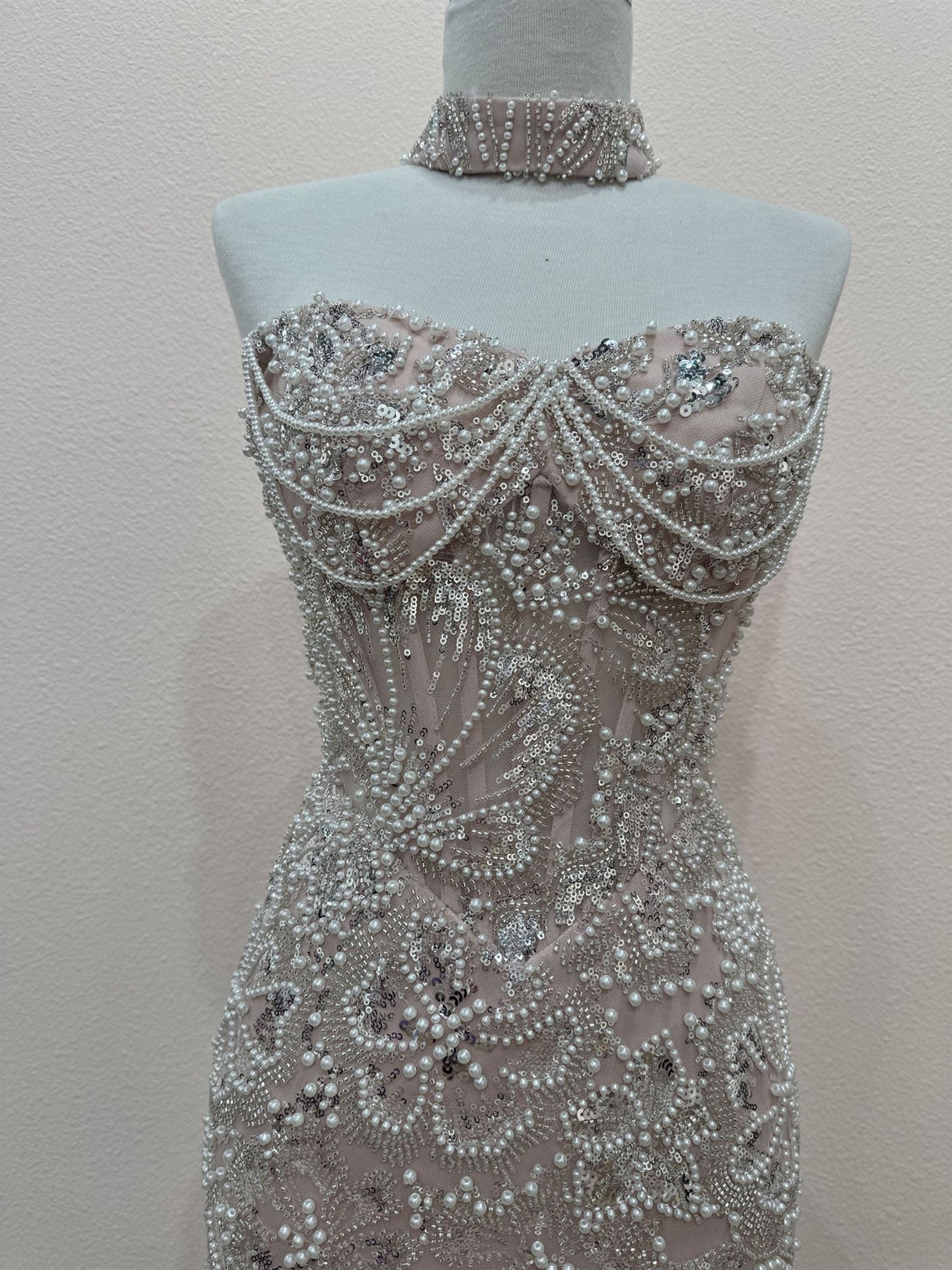 A strapless fitted gown displayed on a mannequin torso, covered in dense beadwork and pearl-like embellishments. The dress has a sheer blush-beige base with silver-toned beading and a matching beaded choker collar.