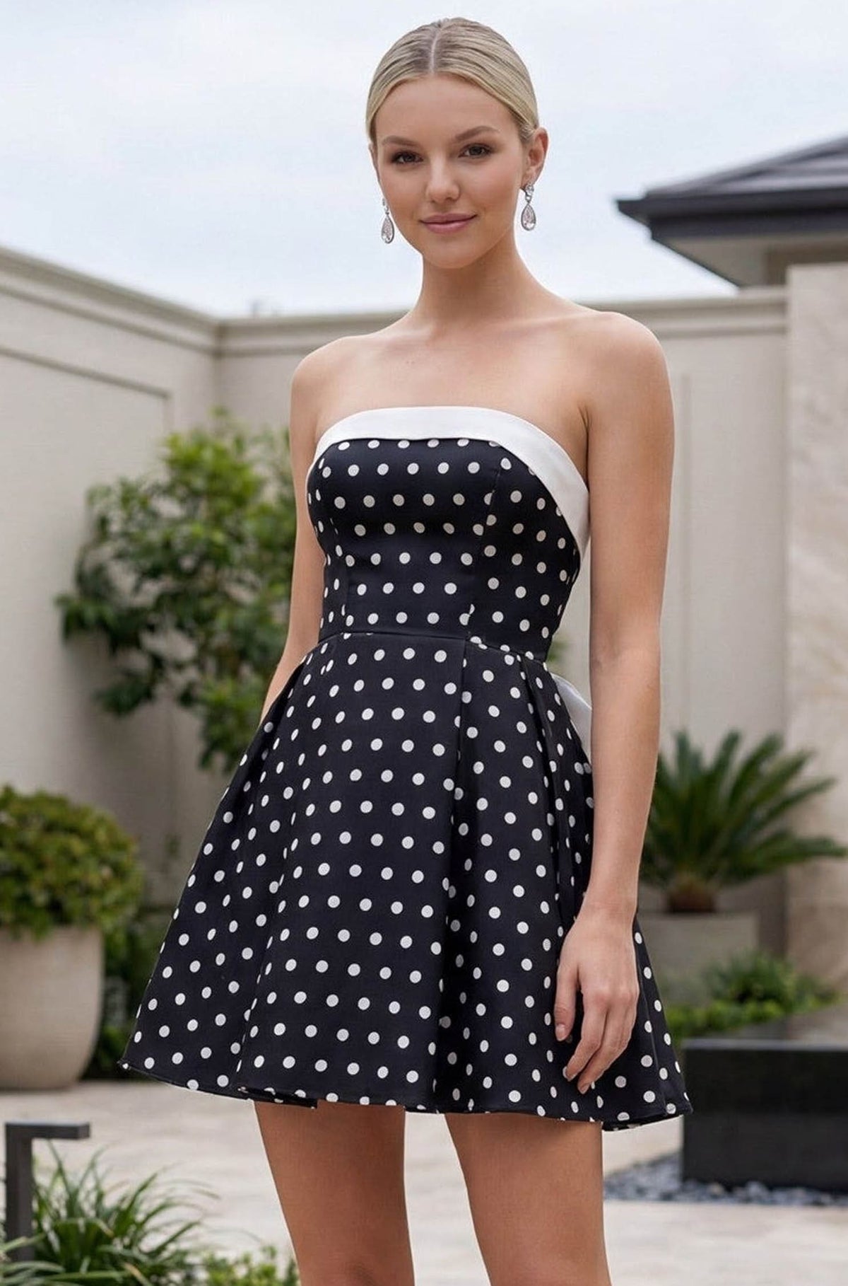 A woman modeling a strapless black dress with white polka dots and a contrasting white band at the neckline, set against an outdoor patio background.