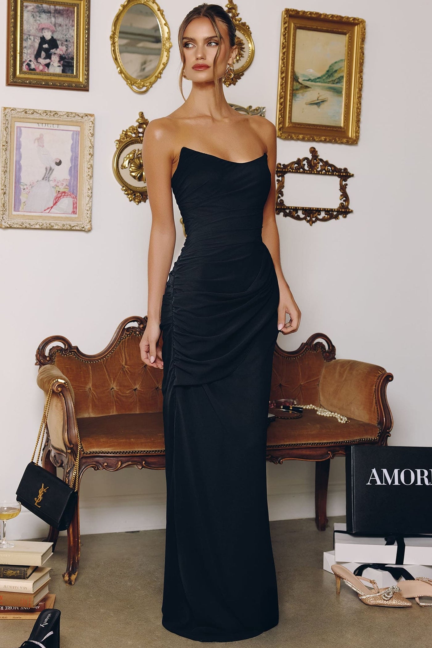 Woman in a black one-shoulder dress standing in an elegant room with a wooden bench and framed pictures.