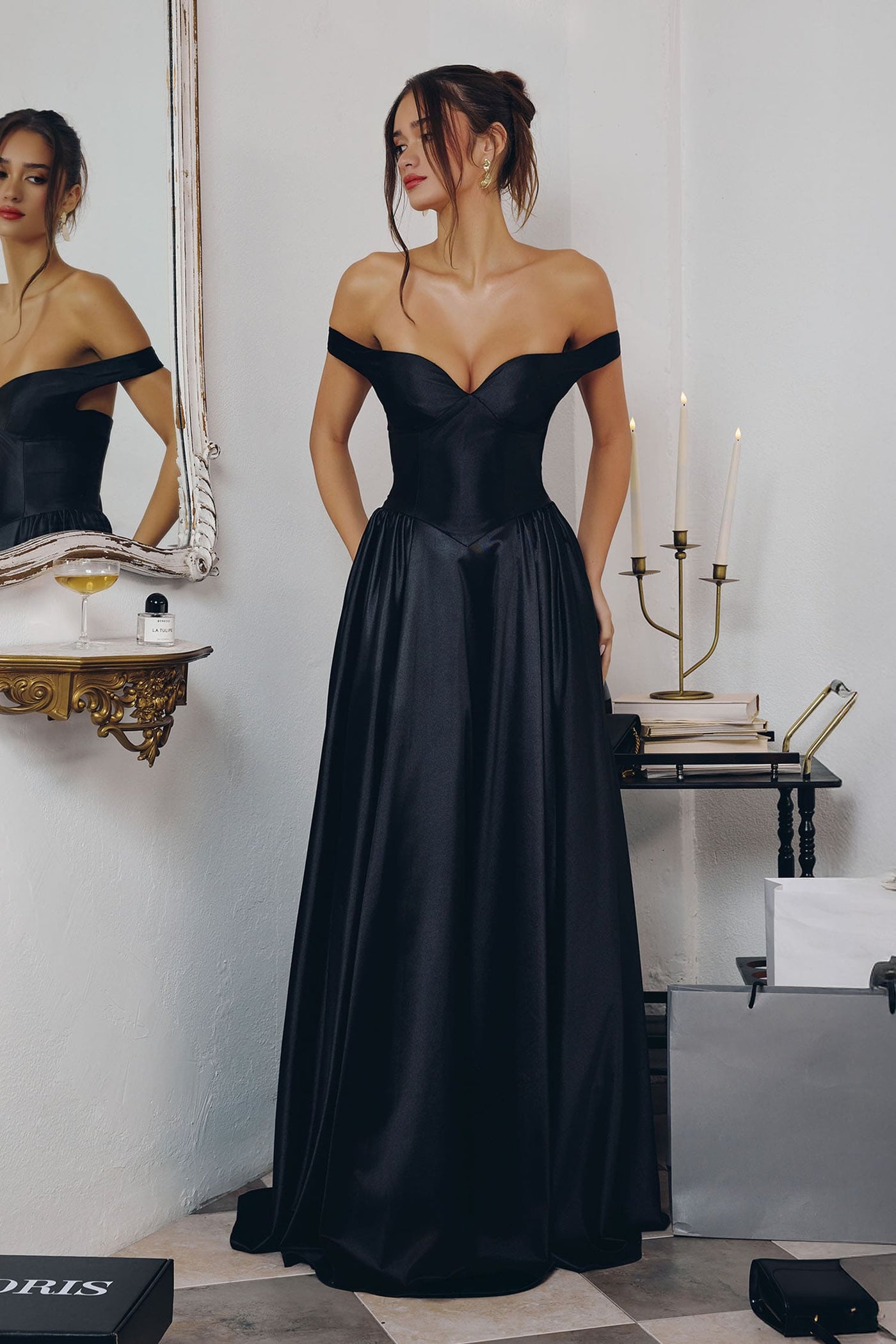 Woman in a black off-shoulder gown standing in an elegant room with candles and a mirror.