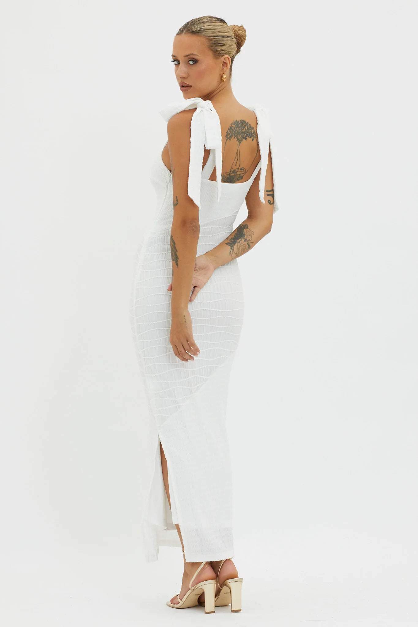 A woman poses gracefully, wearing a form-fitting white dress with tied straps, showcasing intricate tattoos on her back, set against a simple white background. She stands confidently, with one hand at her waist.