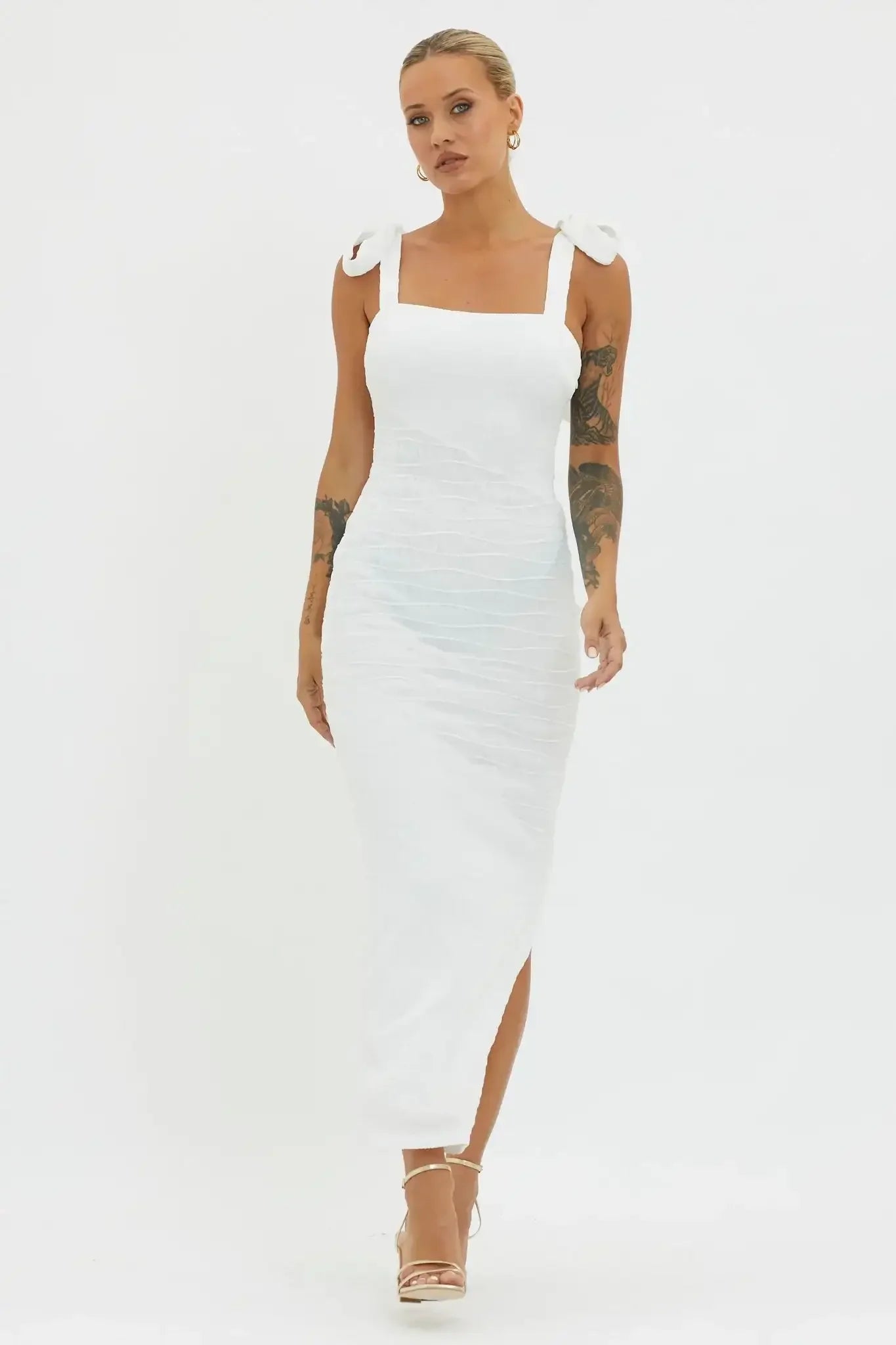 A woman wearing a fitted white dress with bow straps walks confidently, showcasing her tattoos. The neutral background emphasizes her elegant appearance and the dress's textured design.