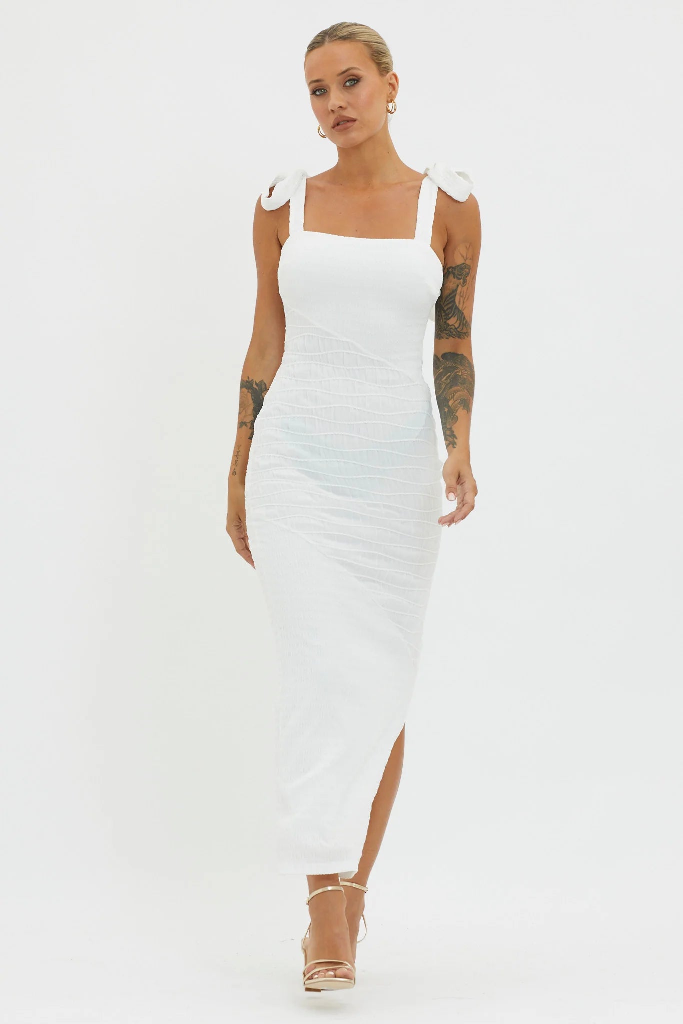 A woman wearing a fitted white dress with bow straps walks confidently, showcasing her tattoos. The neutral background emphasizes her elegant appearance and the dress's textured design.