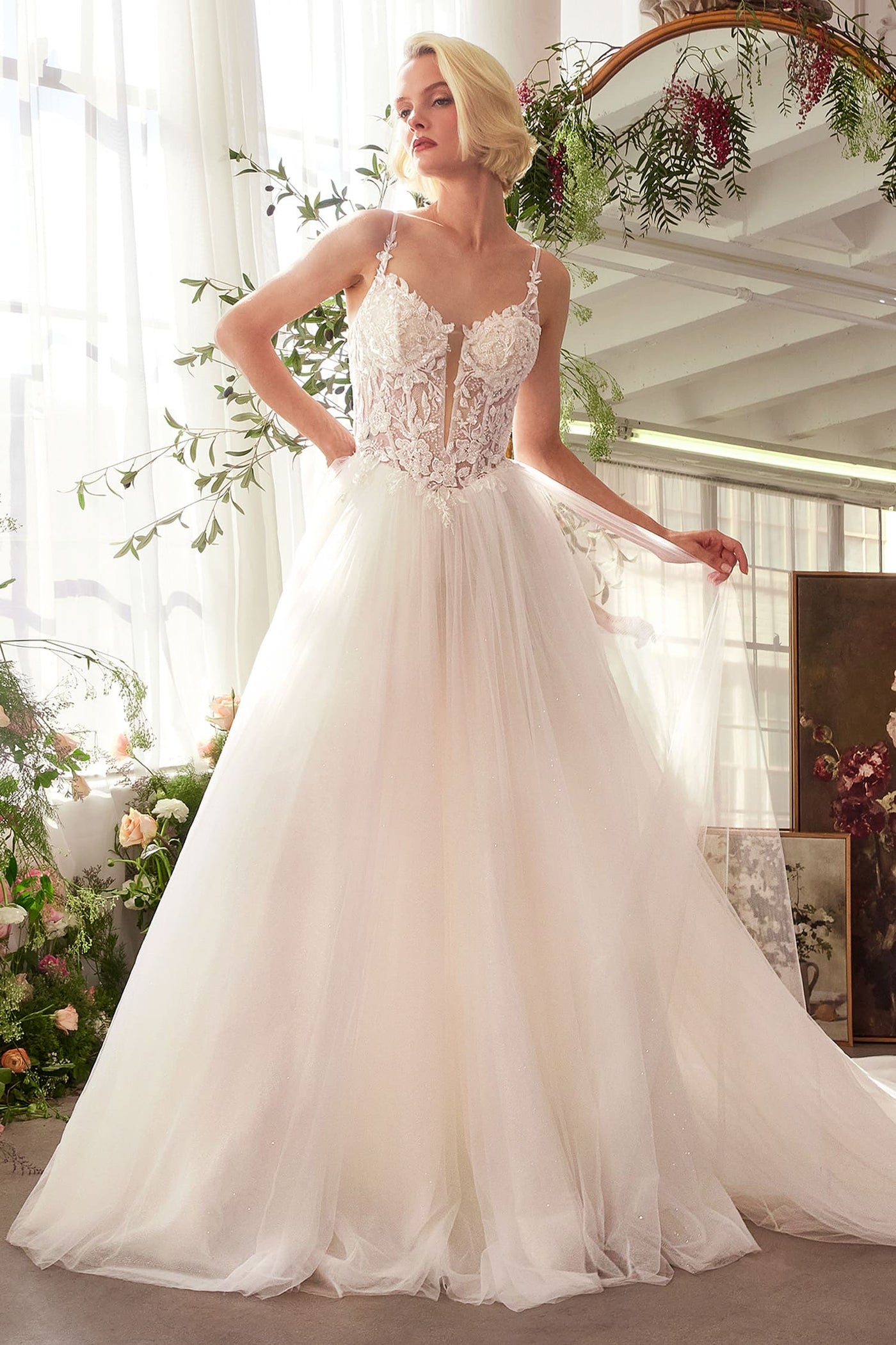 A woman modeling a white wedding gown with a lace bodice, thin straps, and a full tulle skirt in a bright indoor setting with floral decor and a mirror.