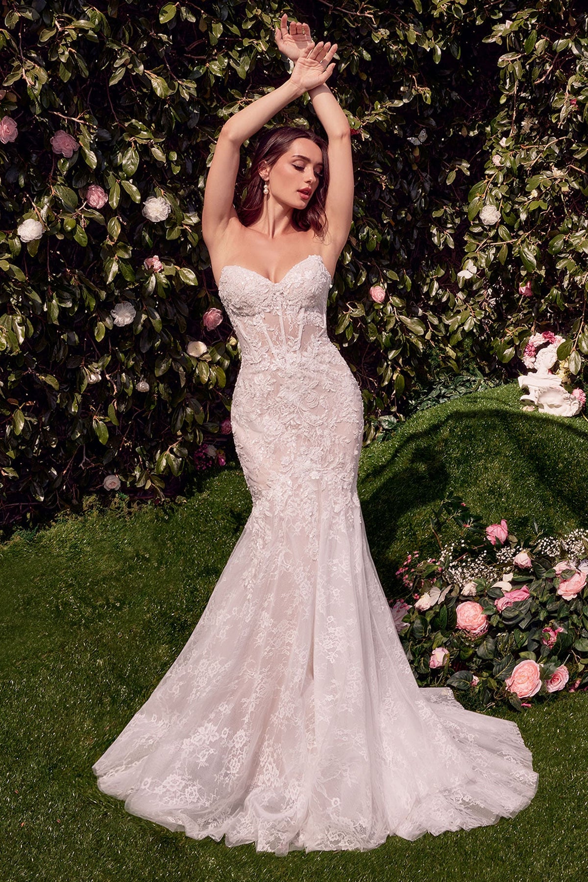 Woman in a white lace wedding dress standing in a garden with flowers and greenery.