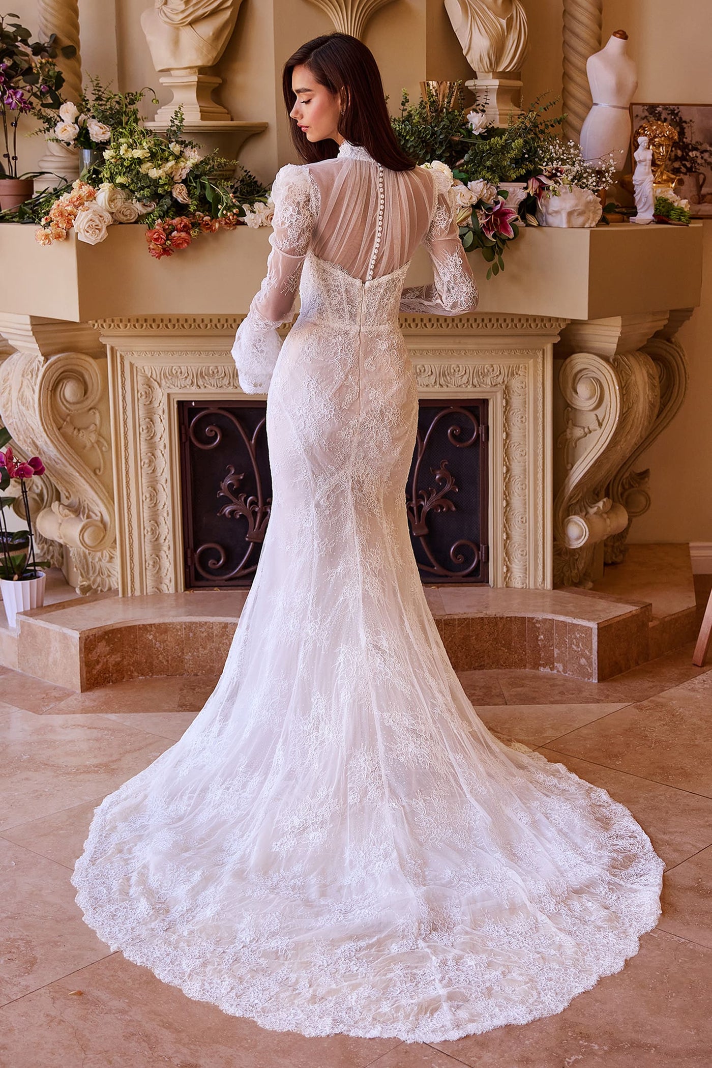 Woman in a white lace wedding dress standing in a decorated room with flowers and a fireplace.
