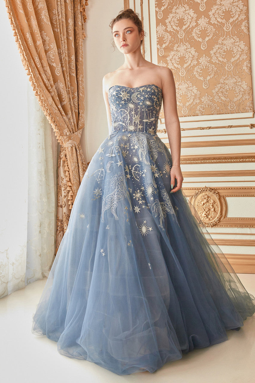 A strapless blue gown adorned with intricate white celestial designs drapes elegantly to the floor. The model stands poised in a lavish room with ornate decor and draped curtains.