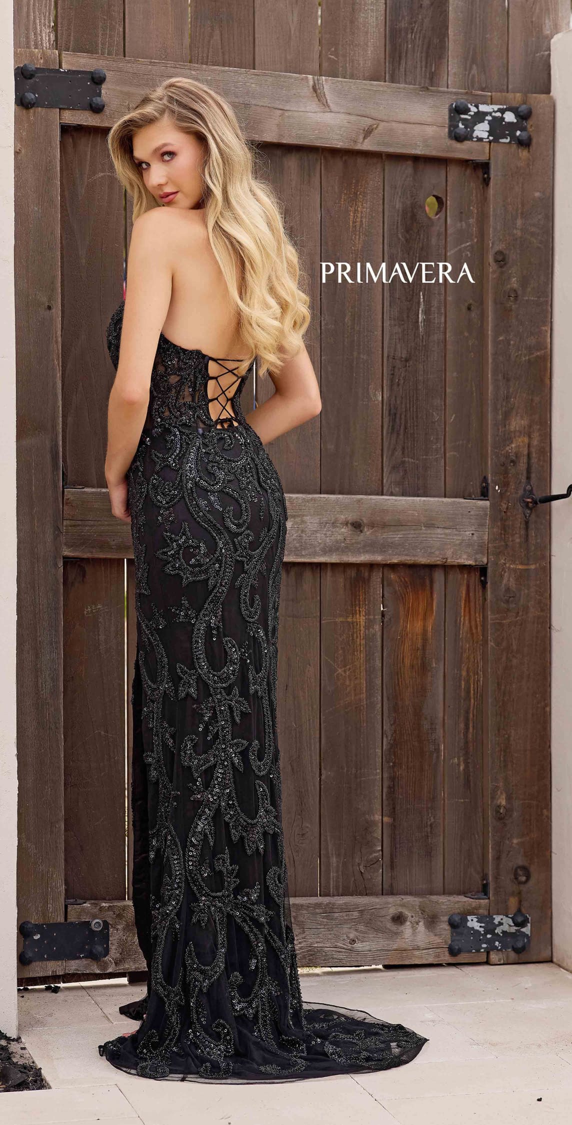 Woman in a black lace dress standing in front of a wooden door with 'Primavera' branding.