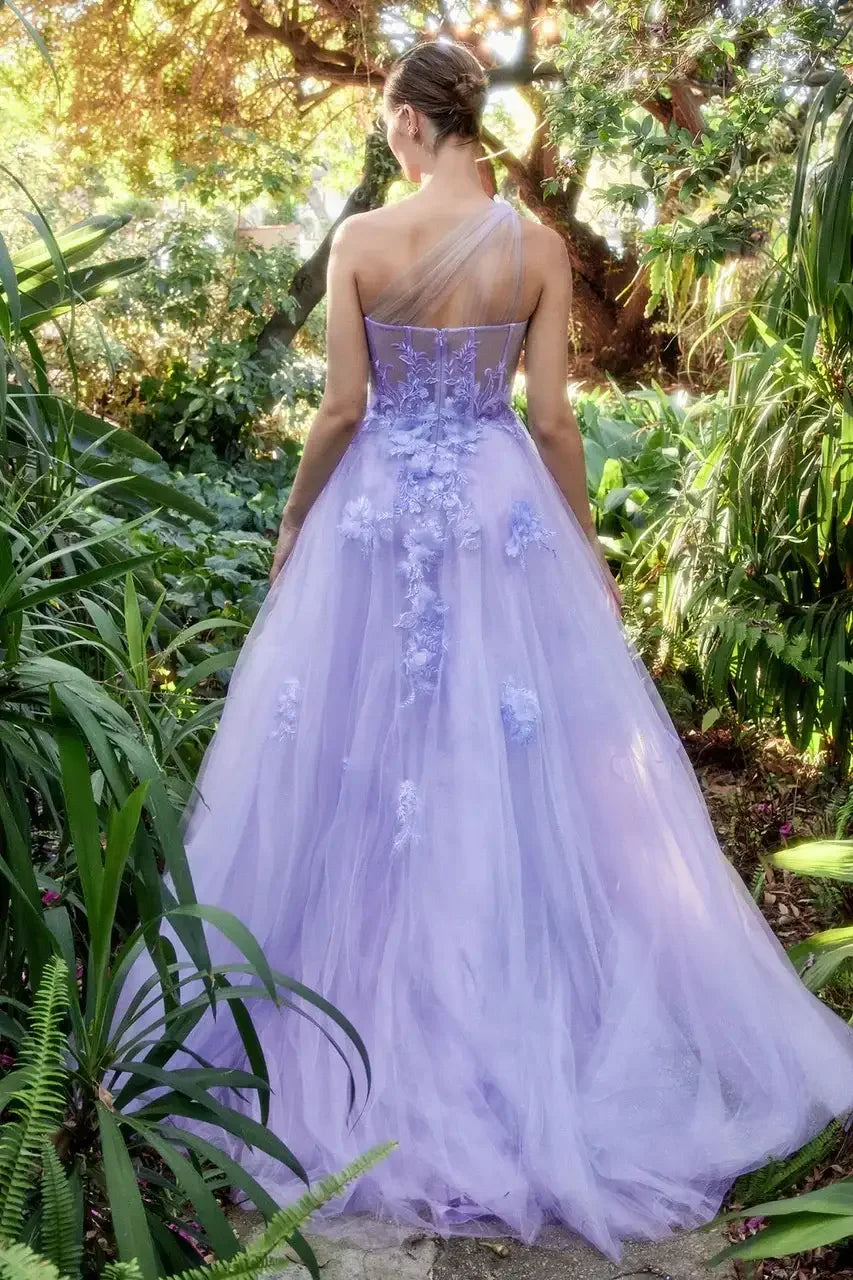 A woman in a lilac, flowing gown adorned with floral embellishments stands facing away, surrounded by lush greenery and dappled sunlight filtering through trees.