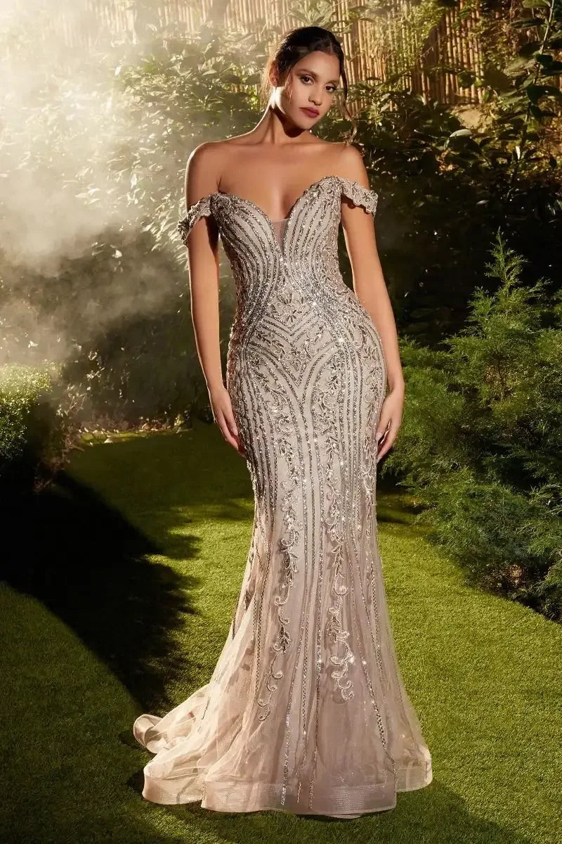 A woman stands elegantly in a sparkly, off-the-shoulder dress. She poses in a lush, green outdoor setting, surrounded by soft mist and foliage, highlighting the garment's intricate details.