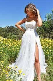 A woman in a flowing white dress stands among vibrant yellow flowers, gently touching her hair while looking to the side, set against a clear blue sky.
