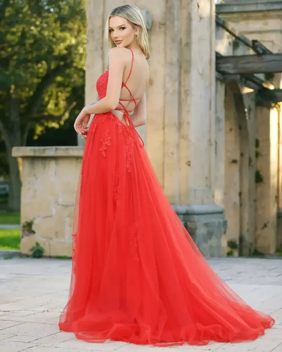 A woman stands in a flowing red gown, showcasing intricate details. She poses gracefully outdoors, with a soft focus on greenery and stone structures in the background.