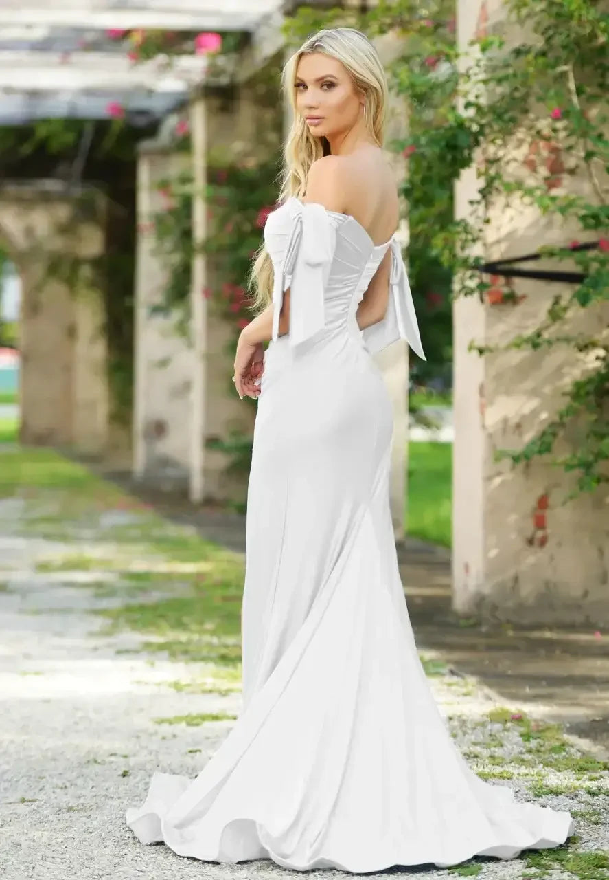 A woman in a fitted white gown stands gracefully, looking back. The setting features greenery and stone pillars, suggesting an outdoor or garden location. Her long hair cascades down her back.