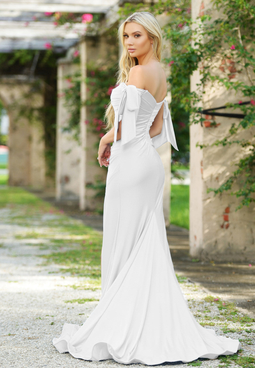 A woman in a fitted white gown stands gracefully, looking back. The setting features greenery and stone pillars, suggesting an outdoor or garden location. Her long hair cascades down her back.