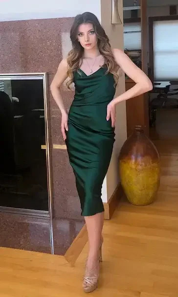 A woman in a green satin dress poses with one hand on her hip and the other slightly raised. She stands in a stylish living room with wooden flooring and a marble fireplace.