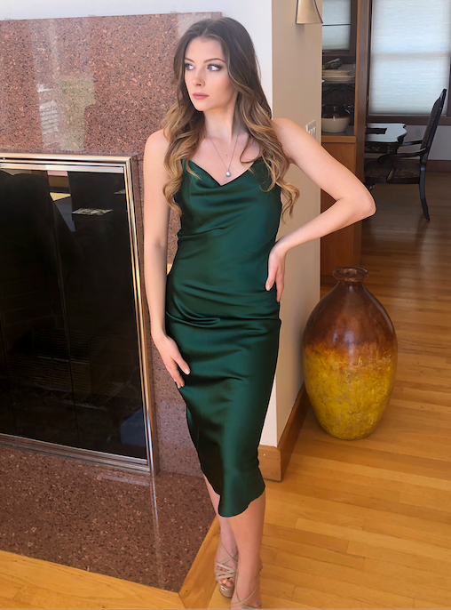 A young woman in a sleek green satin dress stands elegantly next to a modern fireplace, with a large ceramic vase nearby, in a contemporary room with wooden floors and natural light.