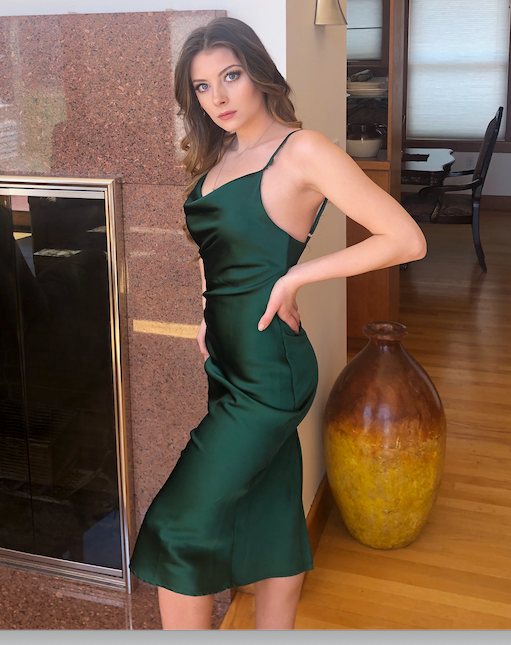A woman in a green satin dress poses with one hand on her hip. She stands beside a modern fireplace in a well-lit, stylish interior featuring wooden flooring and decorative pottery.
