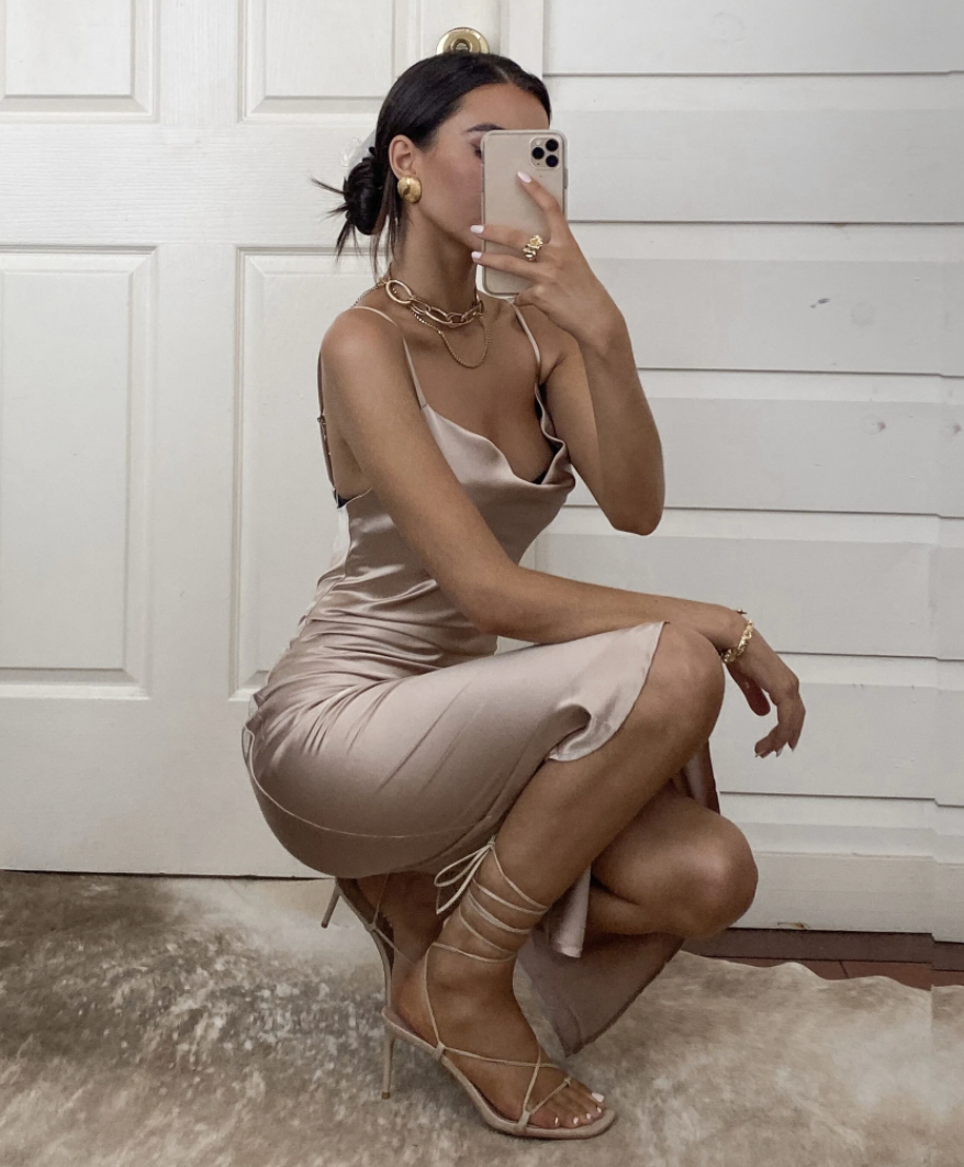 A person in a satin beige dress poses while taking a selfie. They are accessorized with jewelry, sitting on a rug in front of a light-colored door.