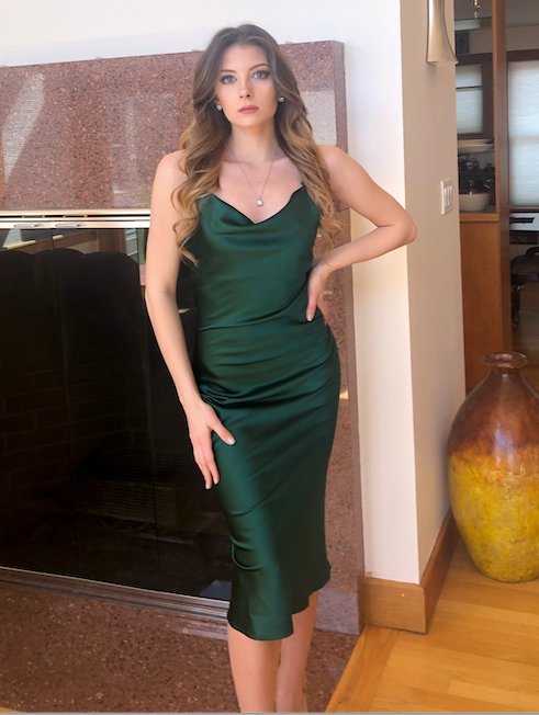 A woman stands confidently in a fitted green satin dress, one hand on her hip, in a well-lit, modern interior with a fireplace and a decorative vase nearby.