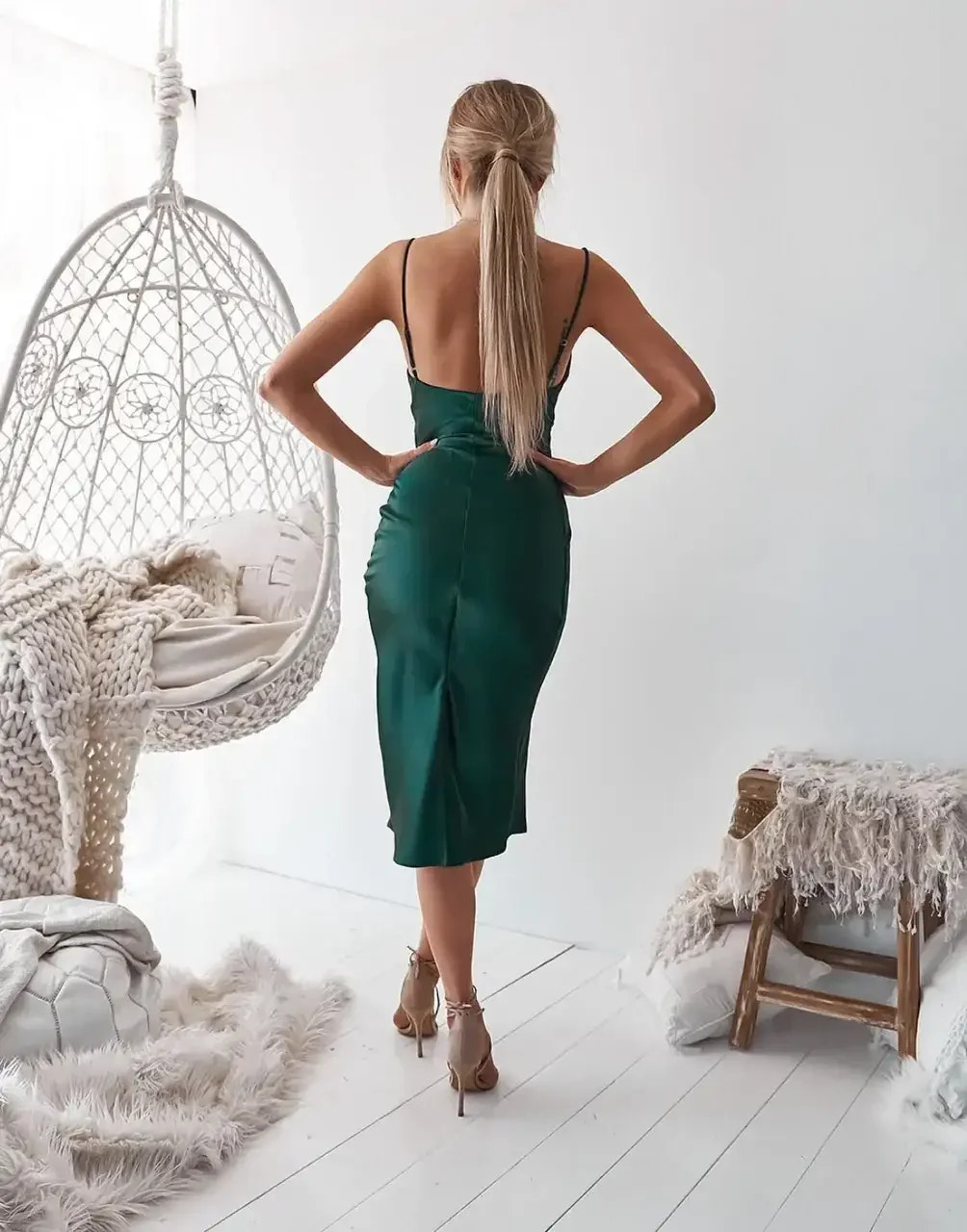 A woman wearing a green satin dress stands gracefully with her back to the viewer. She poses in a bright, neutral room featuring a hanging chair and soft décor.