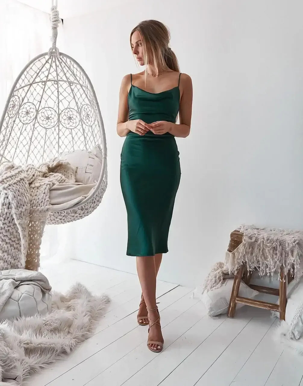 A woman in a green satin dress stands gracefully beside a hanging chair in a bright, minimalistic room. Soft textures and light-colored decor enhance the serene atmosphere.