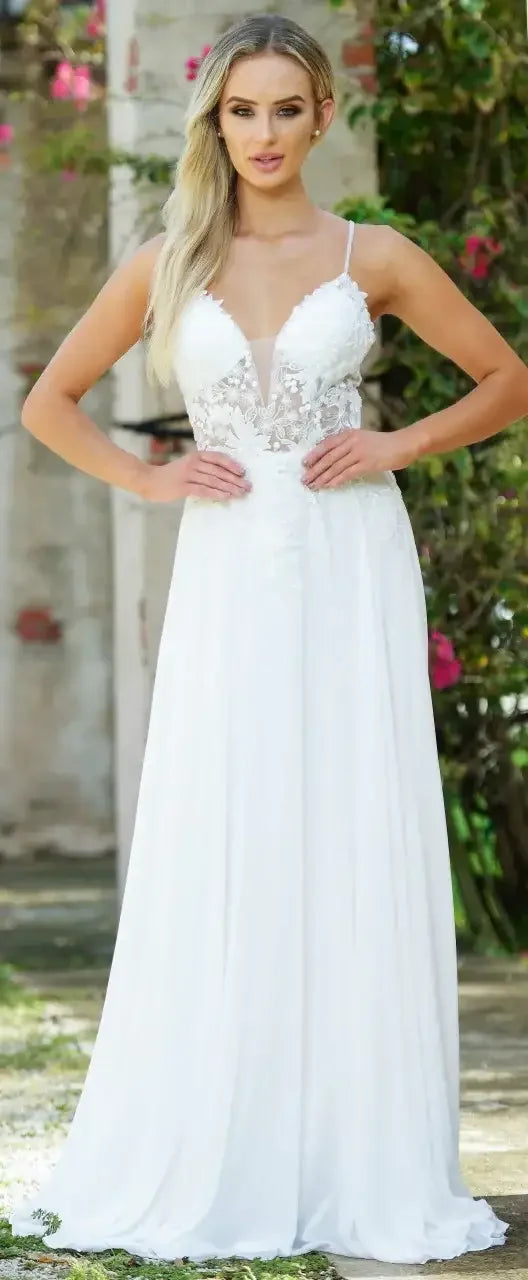 A woman is modeling a flowing white dress with a lace bodice and delicate straps, standing in a garden setting surrounded by greenery and flowers, exuding elegance.