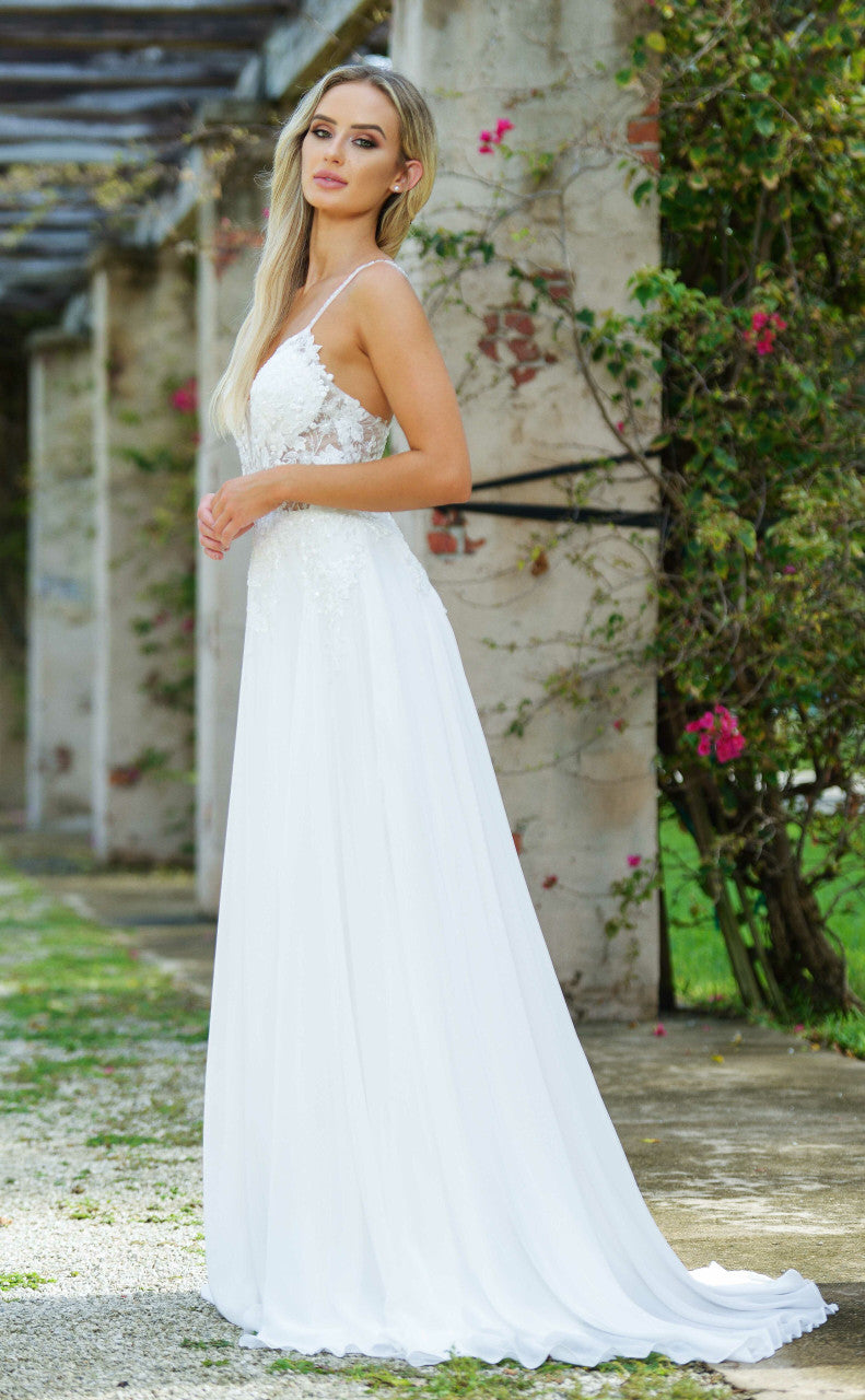 A woman in a flowing white gown stands gracefully outdoors. She poses near weathered stone walls adorned with green vines and pink flowers, surrounded by a lush natural setting.