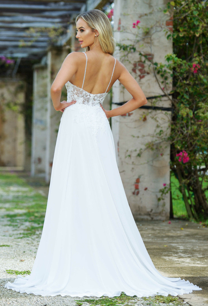 A woman stands in a flowing white dress with delicate lace detailing, showcasing her back. She is positioned outdoors against a backdrop of stone pillars and greenery.