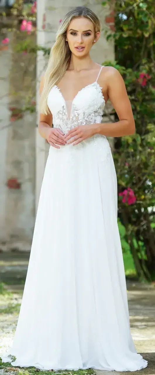 A woman stands gracefully in a flowing, white gown adorned with delicate lace. She holds her hands gently together, surrounded by greenery and textured stone walls, embodying elegance and tranquility.