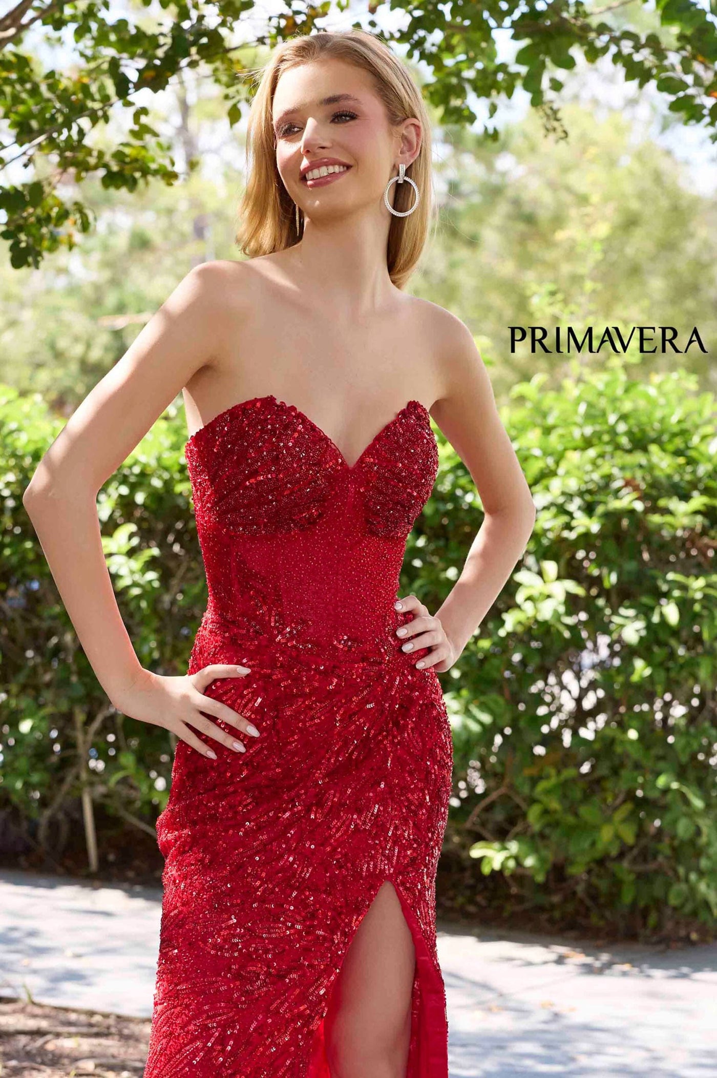 Woman wearing a red dress with beading and a slit, standing outdoors with greenery in the background.