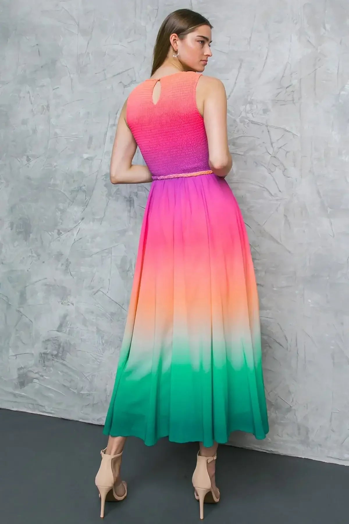 A person stands with their back to the viewer, wearing a vibrant, gradient dress shifting from pink to orange to green. The background features a textured gray wall and dark flooring.