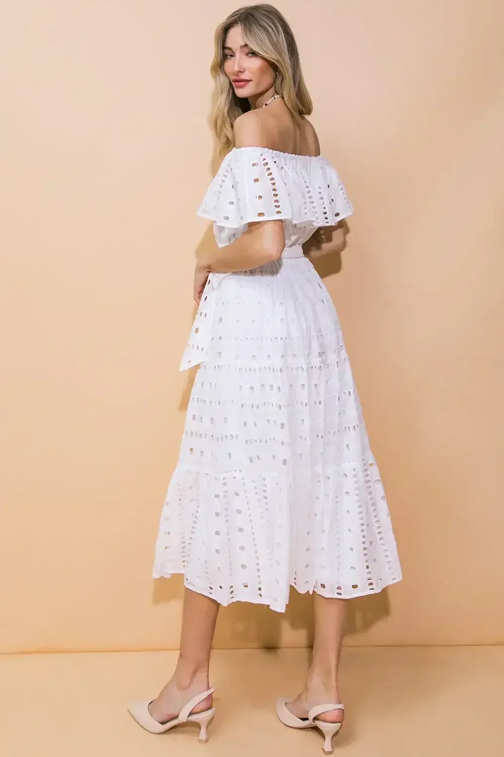 A woman in a white, off-the-shoulder dress stands with her back to the viewer, showing the dress's lace details and a light beige background, exuding a summery, chic vibe.
