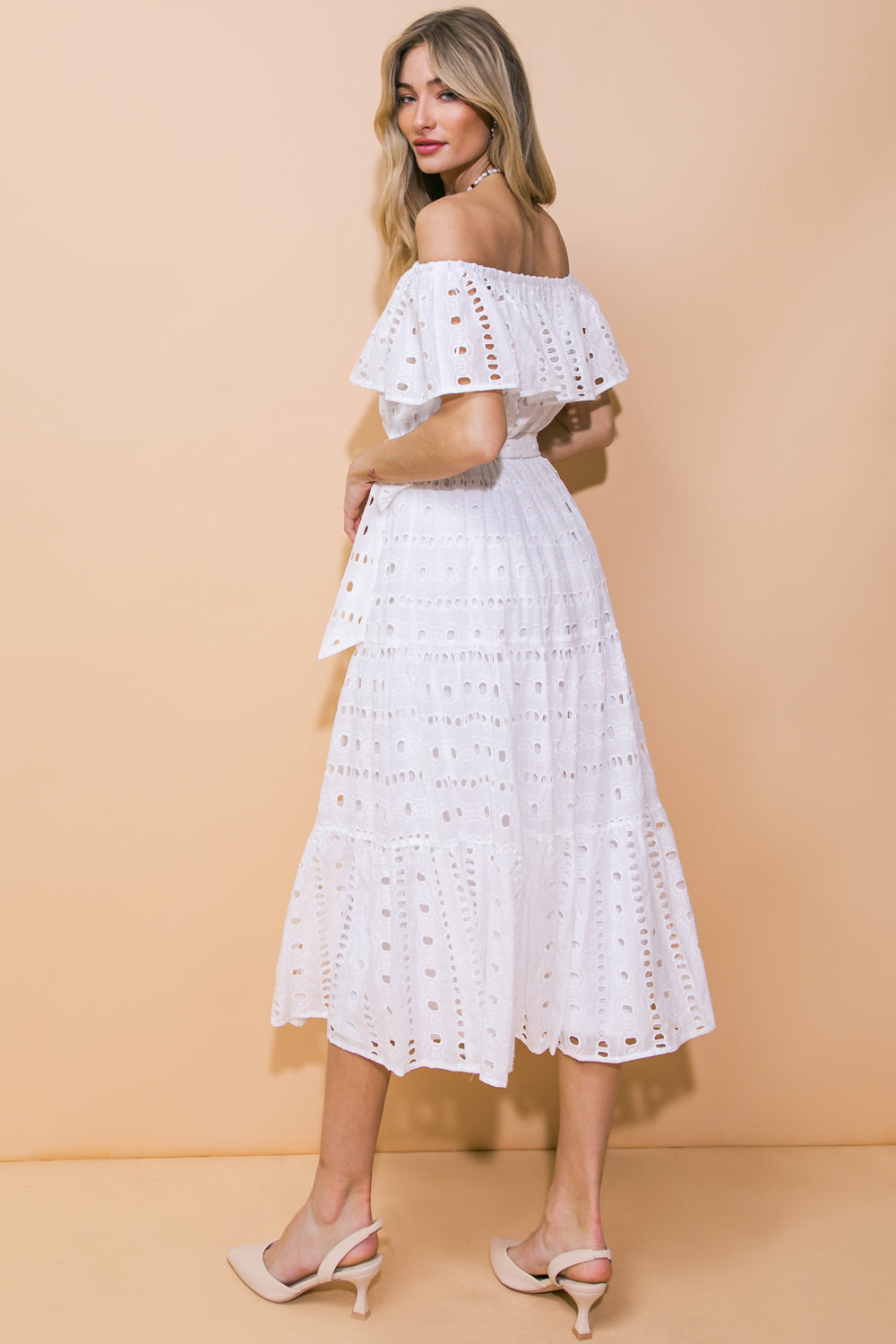 A woman in a white, off-the-shoulder dress stands with her back to the viewer, showing the dress's lace details and a light beige background, exuding a summery, chic vibe.