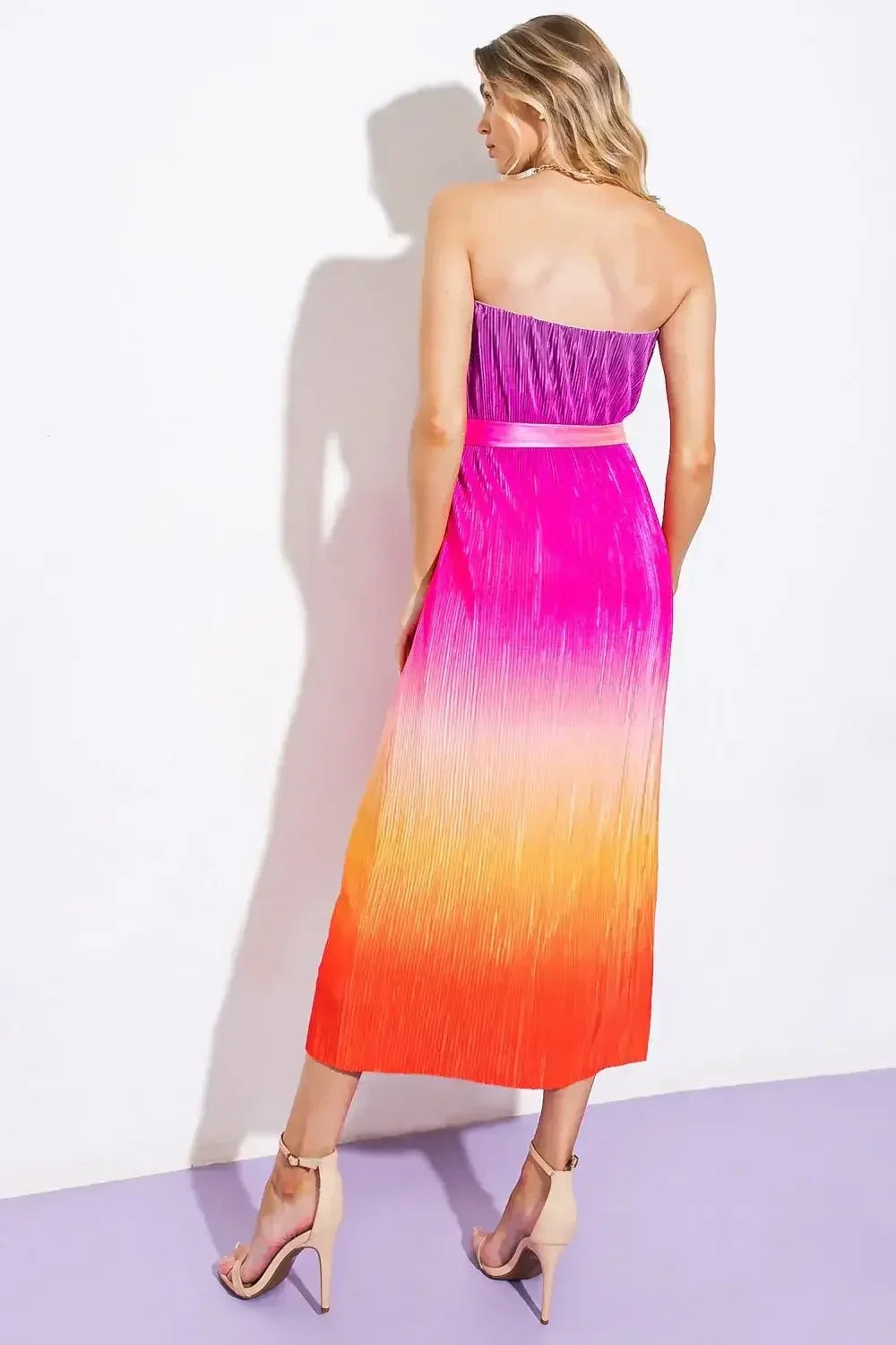 A woman stands with her back turned, wearing a strapless, pleated dress that transitions from purple at the top to orange at the bottom, complemented by beige high-heeled sandals.