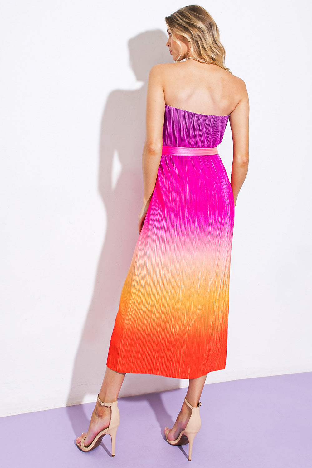 A woman stands with her back turned, wearing a strapless, pleated dress that transitions from purple at the top to orange at the bottom, complemented by beige high-heeled sandals.