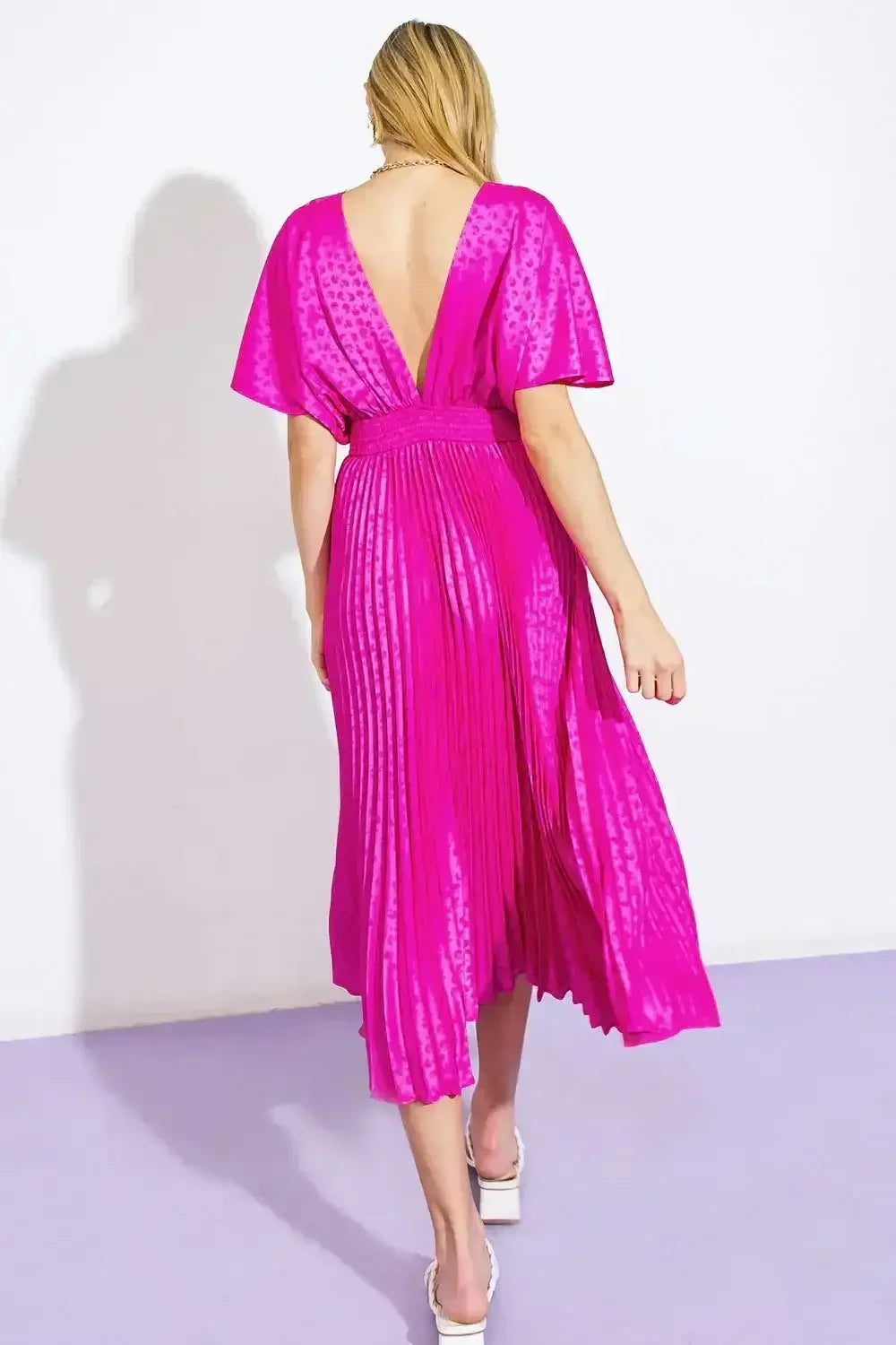 A woman wears a vibrant, pleated pink dress with a deep back in a brightly lit, minimalistic environment, showing her turned away from the viewer on a purple floor.