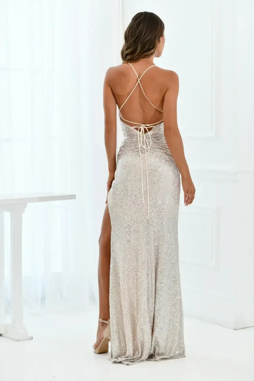 A woman wears a shimmering, silver gown with a low back and crisscross straps. She stands in a bright, airy room with white walls and soft curtains, exuding elegance.